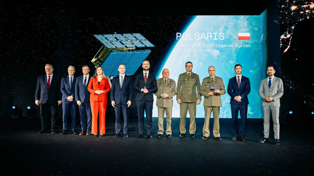 Officials and Polish Army officers on stage at the POLSARIS handover ceremony, with a SAR satellite render and the Polish flag on the screen behind them