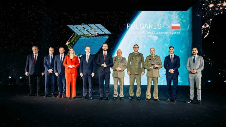 Officials and Polish Army officers on stage at the POLSARIS handover ceremony, with a SAR satellite render and the Polish flag on the screen behind them